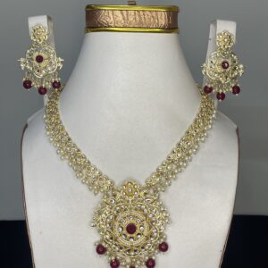 Gold-tone Kundan bridal necklace set with ruby red stones, pearls, and matching earrings displayed on a jewelry stand.