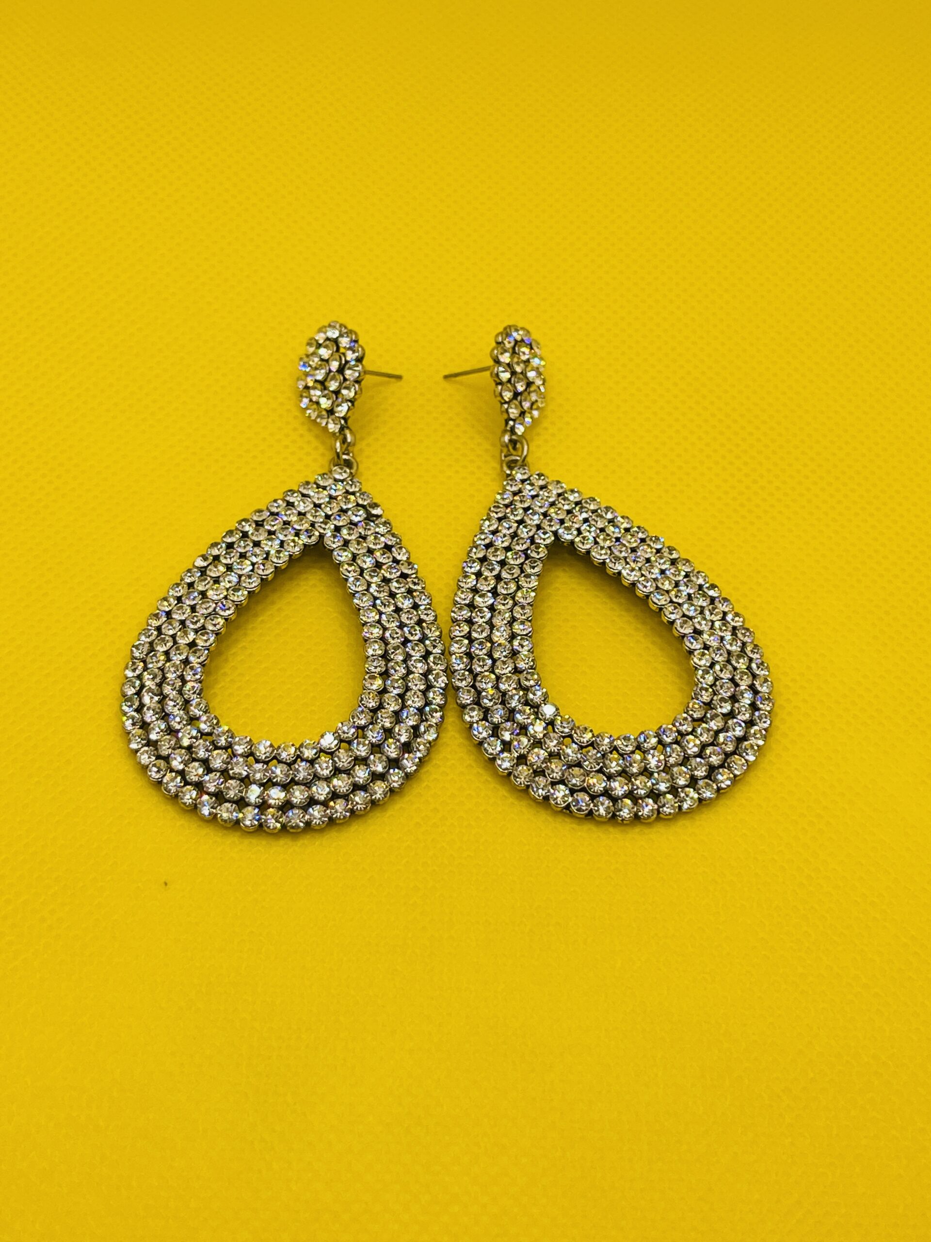 Elegant crystal teardrop earrings with multi-row design on yellow background Sparkling teardrop statement earring displayed on white holder with crystals Pair of crystal teardrop statement earrings laid flat on yellow background